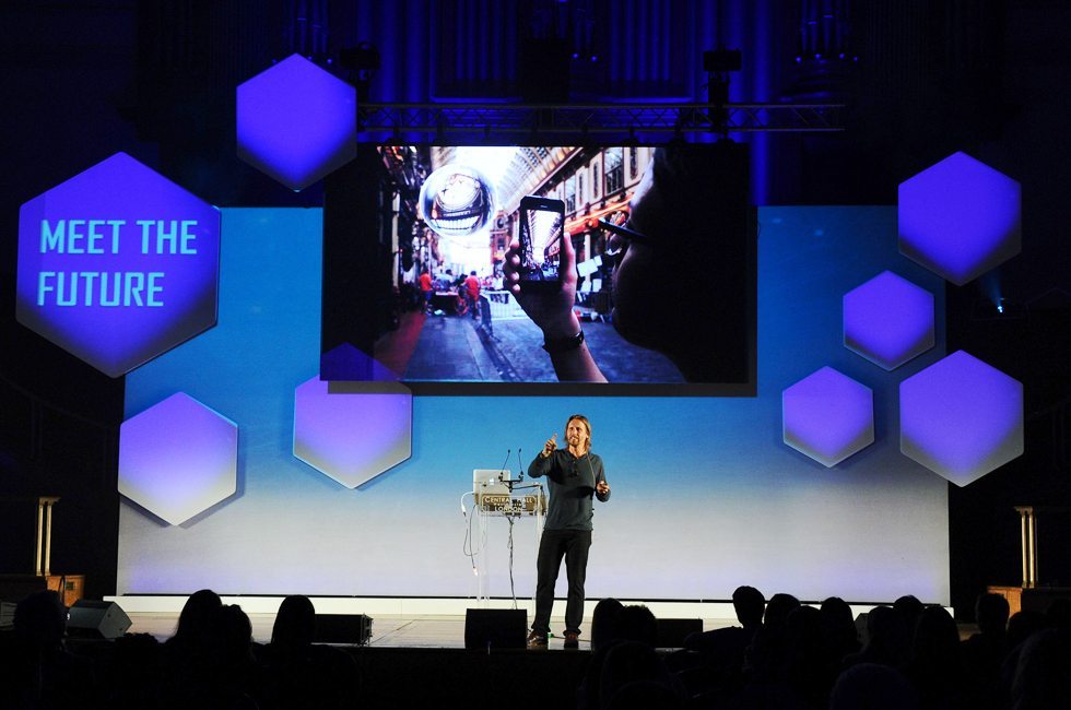 Presenter on stage at technology seminar, under screen playing video content